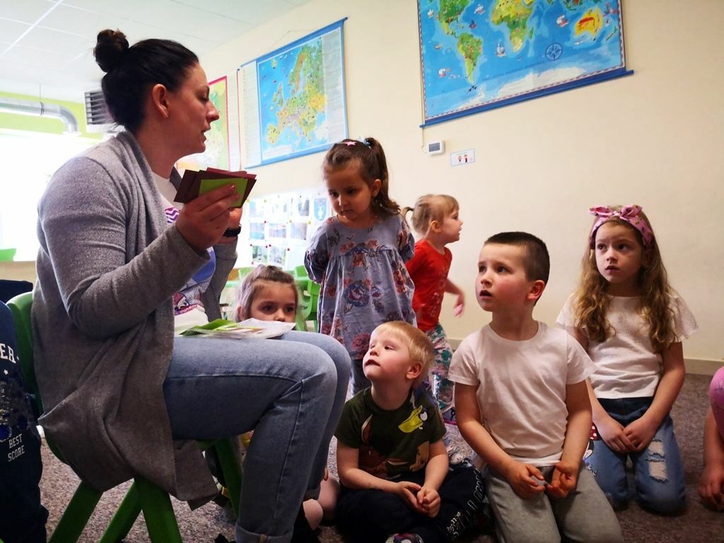 na zdjęciu widać bibliotekarkę czytającą z kolorowych karteczek siedzące na małym krześle oraz słuchające ją dzieci