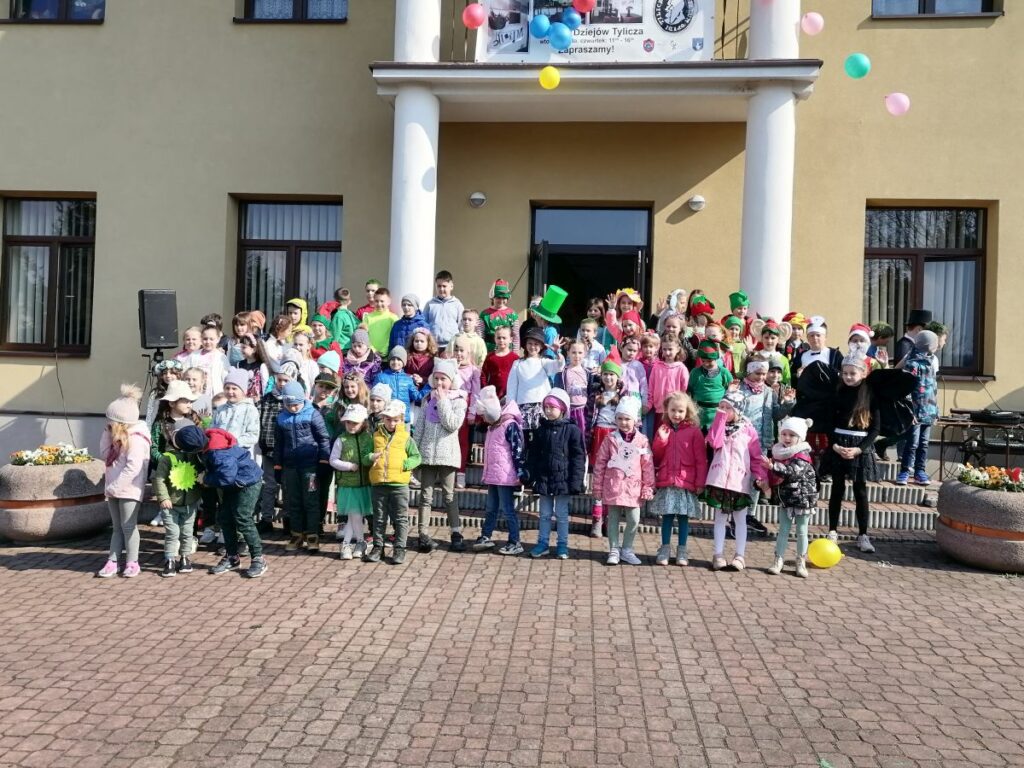 Na zdjęciu widoczna jest grupa ok 81 uczniów z Kl. I – III Szkoły Podstawowej w Tyliczu i Zespołu Szkolno-Przedszkolnego w Muszynce, uczestniczących w imprezie plenerowej zorganizowanej przez Bibliotekę Publiczną, Świetlicę „Przystań” i Centrum Kultury. Dzieci przebrane są w kolorowe stroje m.in. za elfy, krasnale, wróżki wpisując się w motyw przewodni imprezy. Uczniowie pozują do wspólnego zdjęcia przed budynkiem WDK w Tyliczu.