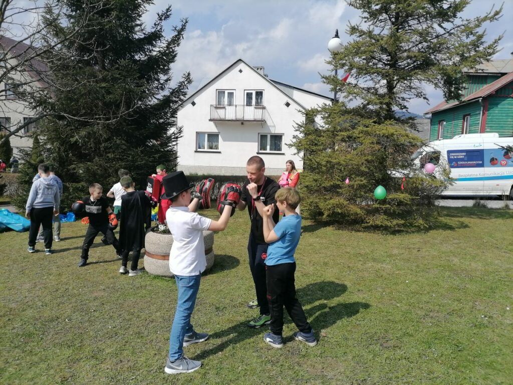 Na zdjęciu widoczny jest trener kickboxingu i grupka chłopców podczas treningu. Dzieci maja ubrane rękawice bokserskie i trzymają tarcze do kickboxingu. W tle drzewa, domy i bus.