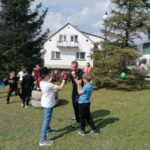 Na zdjęciu widoczny jest trener kickboxingu i grupka chłopców podczas treningu. Dzieci maja ubrane rękawice bokserskie i trzymają tarcze do kickboxingu. W tle drzewa, domy i bus.