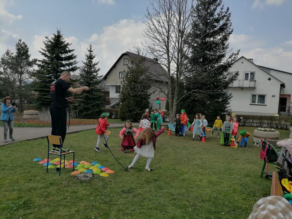 Na zdjęciu widoczny jest animator, który stoi na krześle i kręci wokół siebie liną. Wokół krzesła na którym stoi animator ułożone są kolorowe pachołki. Zdjęcie pokazuje zabawę podczas której grupa dzieci próbuje zdobyć kolorowe pachołki tak by nie dotknęła ich lina kręcona przez animatora.