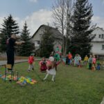 Na zdjęciu widoczny jest animator, który stoi na krześle i kręci wokół siebie liną. Wokół krzesła na którym stoi animator ułożone są kolorowe pachołki. Zdjęcie pokazuje zabawę podczas której grupa dzieci próbuje zdobyć kolorowe pachołki tak by nie dotknęła ich lina kręcona przez animatora.