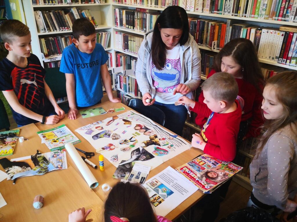 na zdjęciu widoczna jest sala biblioteczna, regały z książkami, na środku sali stoi stół, przy stole stoją dzieci i bibliotekarz, wykonują pracę plastyczną
