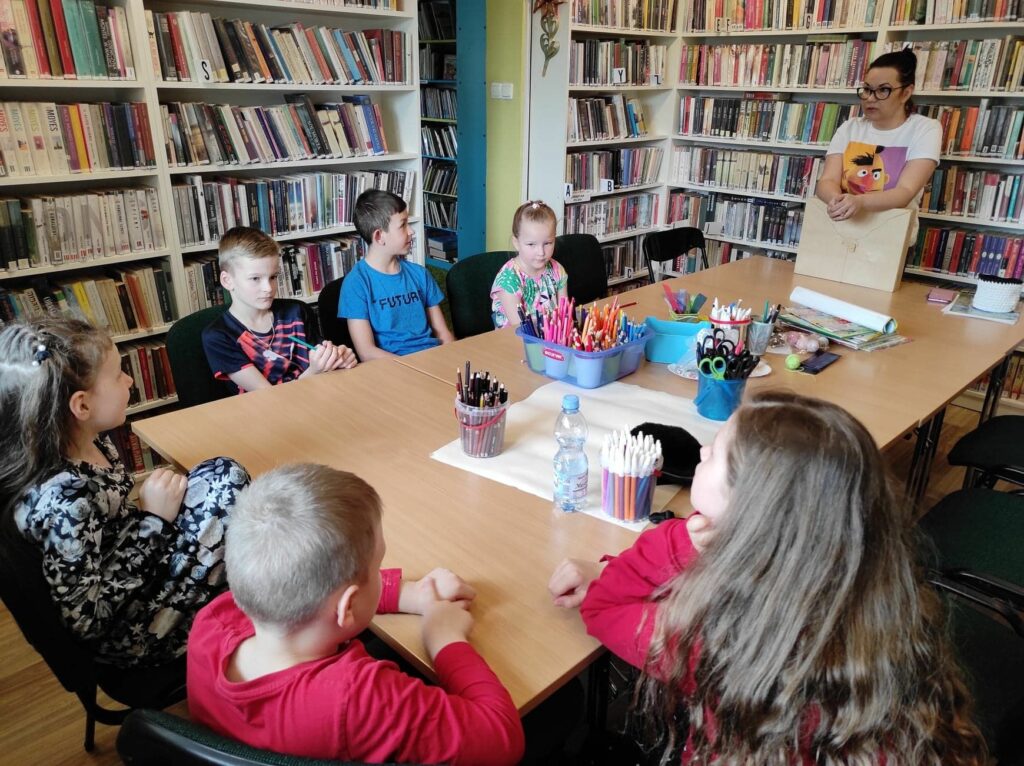 na zdjęciu widoczna jest sala biblioteczna, regały z książkami, na środku sali stoi stół, wokół stołu gromadzą się dzieci i bibliotekarz