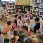 na zdjęciu widać dzieci siedzące na podłodze oraz bibliotekarkę czytającą im książki.