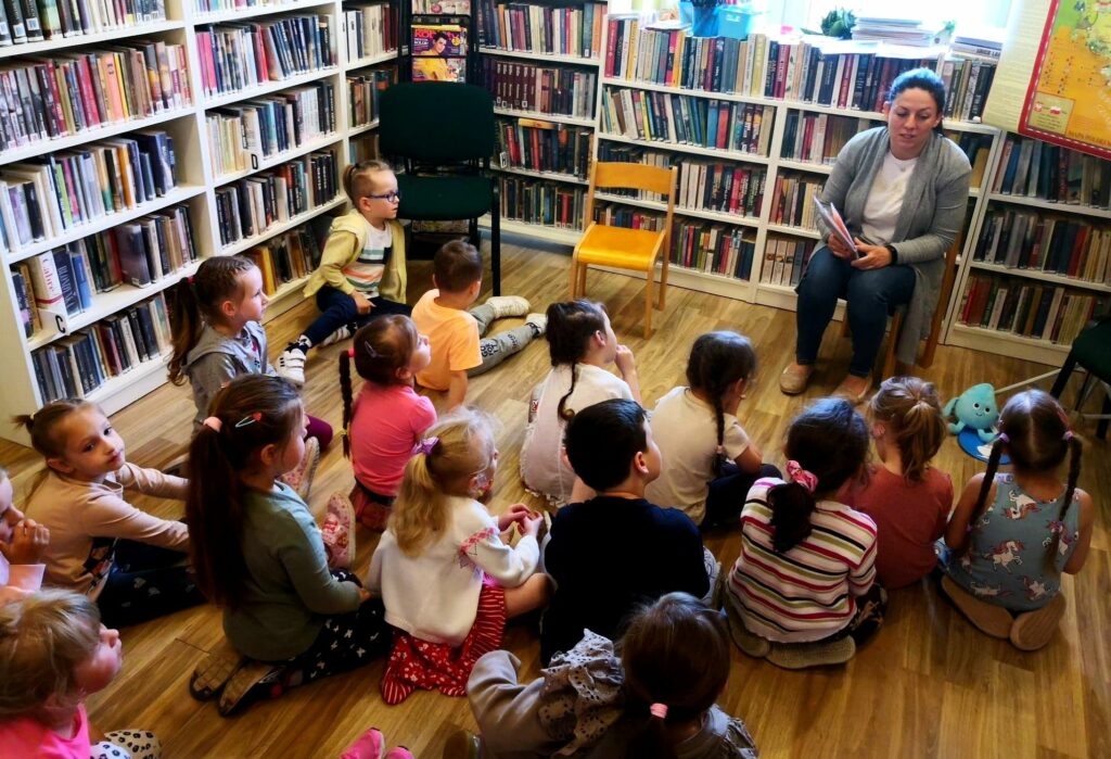 na zdjęciu widać bibliotekarkę siedząca na krzesełku a przed nią siedzące dzieci w wieku przedszkolnym.