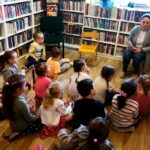 na zdjęciu widać bibliotekarkę siedząca na krzesełku a przed nią siedzące dzieci w wieku przedszkolnym.