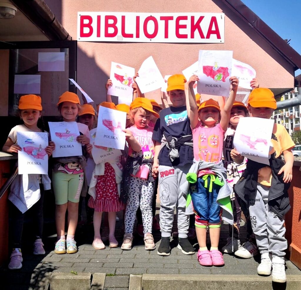 na zdjęciu widać dzieci w pomarańczowych czapeczkach z daszkiem stojące przed biblioteką. Dzieci są w wieku przedszkolnym i trzymają w rękach pomalowane przez siebie ilustracje przedstawiające mapę Polski.