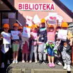 na zdjęciu widać dzieci w pomarańczowych czapeczkach z daszkiem stojące przed biblioteką. Dzieci są w wieku przedszkolnym i trzymają w rękach pomalowane przez siebie ilustracje przedstawiające mapę Polski.
