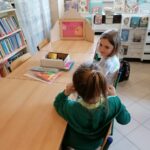 Przy bibliotecznym stole siedzą biorące udział w spotkaniu dziewczynki i oglądają teatrzyk kamishibai pt. „Mój przyjaciel Kemushi”.