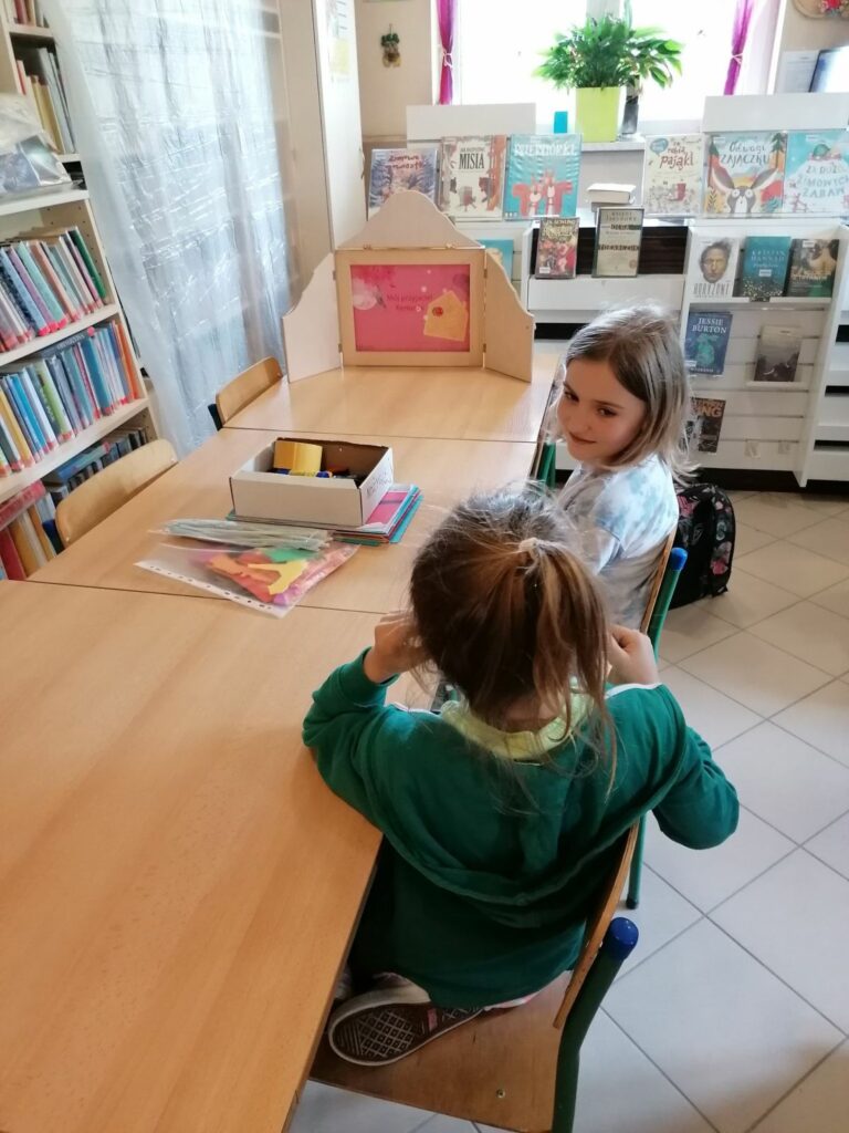Przy bibliotecznym stole siedzą biorące udział w spotkaniu dziewczynki i oglądają teatrzyk kamishibai pt. „Mój przyjaciel Kemushi”.