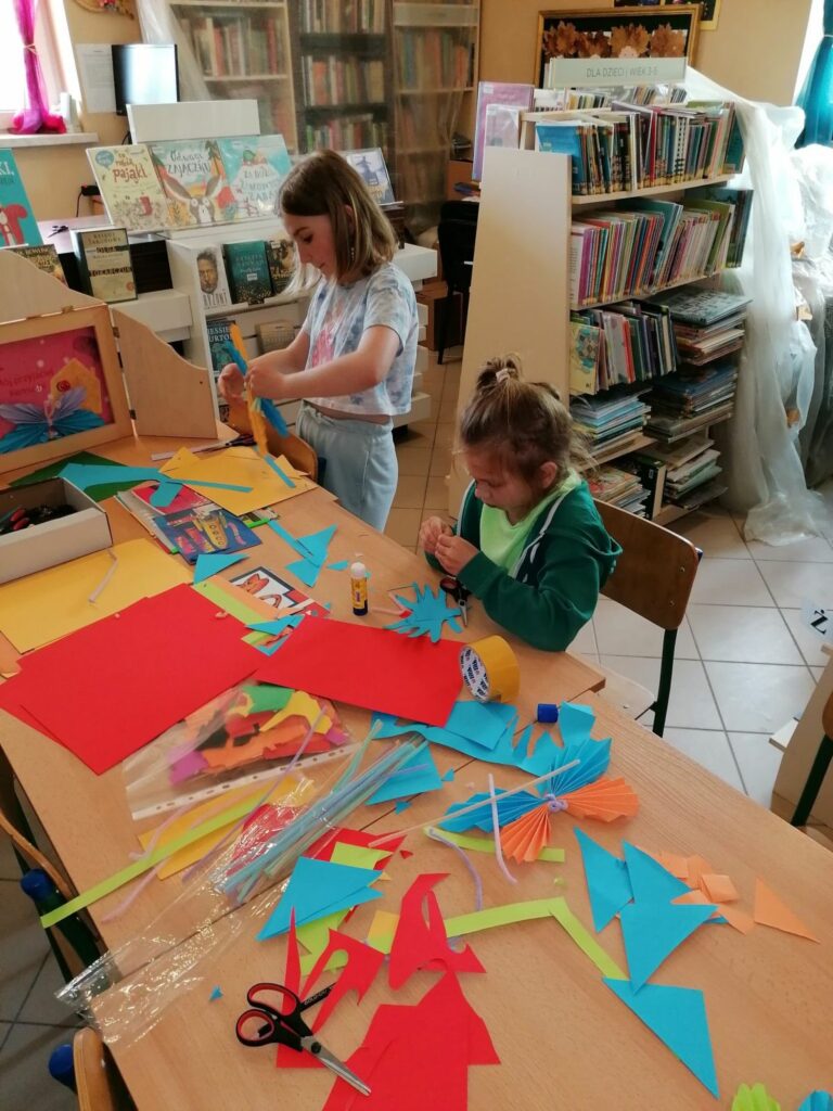 Dzieci pracują przy zapełnionym materiałami plastycznymi stole wycinając i klejąc swoje piękne papierowe motyle.