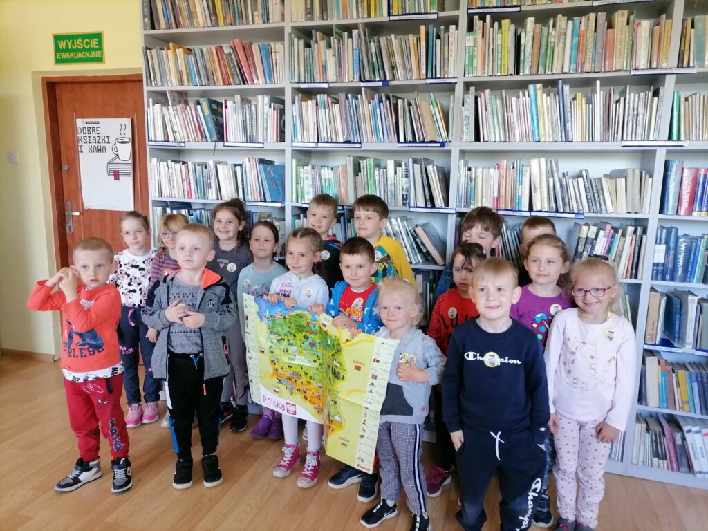 Na zdjęciu widoczna jest grupa 17 dzieci (8 chłopców i 9 dziewczynek) uczestniczących w zajęciach w bibliotece pozujących na tle regałów z książkami. Troje dzieci trzyma mapę Polski.