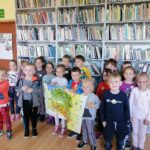 Na zdjęciu widoczna jest grupa 17 dzieci (8 chłopców i 9 dziewczynek) uczestniczących w zajęciach w bibliotece pozujących na tle regałów z książkami. Troje dzieci trzyma mapę Polski.