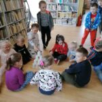 Na zdjęciu widoczna jest grupa 13 przedszkolaków (7 chłopców i 6 dziewczynek) układających "matematyczny szczyt" z kubeczków plastikowych podczas zajęć zorganizowanych w bibliotece. W tle regały z książkami.
