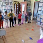 Na zdjęciu widoczna jest grupa 8 przedszkolaków (2 chłopców i 6 dziewczynek) uczestniczących w zabawie "łowienie rybek" podczas zorganizowanych zajęć w bibliotece. W tle regały z książkami.