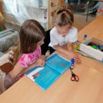 Przy stple siedzą dwie dziewczynki i ogldają książeczkę
