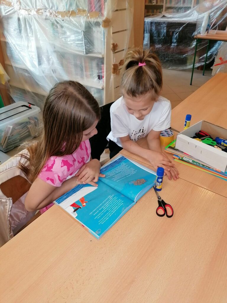 Przy stple siedzą dwie dziewczynki i ogldają książeczkę