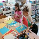 Przy stole stoją dwie dziewczynki w wieku szkolnym i wykonują pracę plastyczna laurkę na dzień Mamy