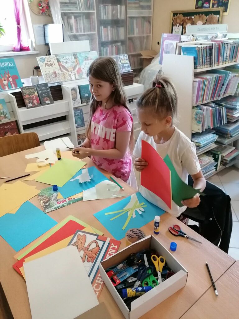Przy stole stoją dwie dziewczynki w wieku szkolnym i wykonują pracę plastyczna laurkę na dzień Mamy