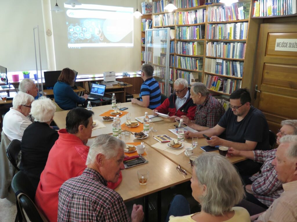 W sali czytelni bibliteki pod Górą Parkową przy dużym stole siedzi siedmiu mążczyzn i cztery kobiety. Na ścianie wyświetkane sa slajdy ze starymi zdjęciami Krynicy.