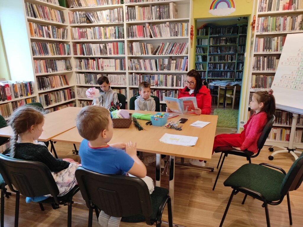 na zdjęciu widać salę biblioteczną, regały z książkami, na środku sali stoi stół, wokół stołu siedzą dzieci i bibliotekarz, bibliotekarz czyta książkę