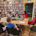 na zdjęciu widać salę biblioteczną, regały z książkami, na środku sali stoi stół, wokół stołu siedzą dzieci i bibliotekarz, bibliotekarz czyta książkę