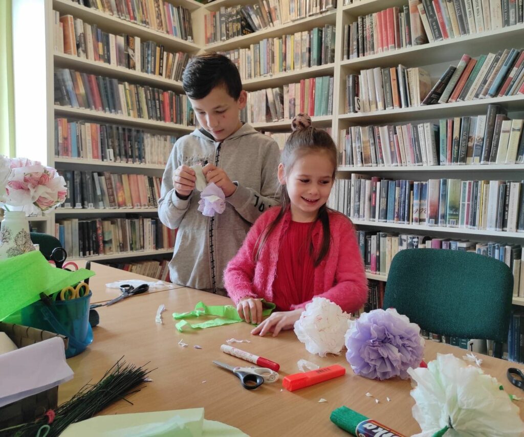 na zdjęciu widać salę biblioteczną, regały z książkami, stół, przy stole stoi chłopiec i dziewczynka, wykonują pracę plastyczną