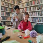 na zdjęciu widać salę biblioteczną, regały z książkami, stół, przy stole stoi chłopiec i dziewczynka, wykonują pracę plastyczną