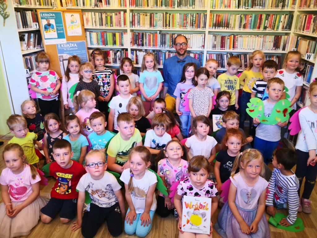 Bardzo duża grupa dzieci stoi do zdjęcia. W środku Pan poeta. Z tyłu za nimi regały biblioteczne z na nich książki. Niektóre z dzieci trzymają w reku książki autorsta pana poety.
