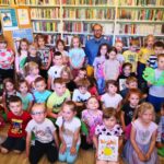 Bardzo duża grupa dzieci stoi do zdjęcia. W środku Pan poeta. Z tyłu za nimi regały biblioteczne z na nich książki. Niektóre z dzieci trzymają w reku książki autorsta pana poety.