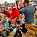 W sali bibliotecznej na ziemi siedzą dzieci po środku klęczy Pan Poeta. Pan Poeta w reku trzyma dużą maskotkę