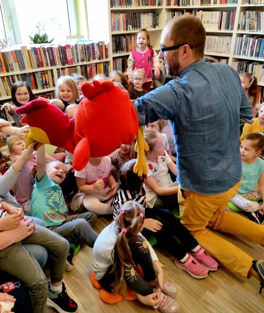 W sali bibliotecznej na ziemi siedzą dzieci po środku klęczy Pan Poeta. Pan Poeta w reku trzyma dużą maskotkę