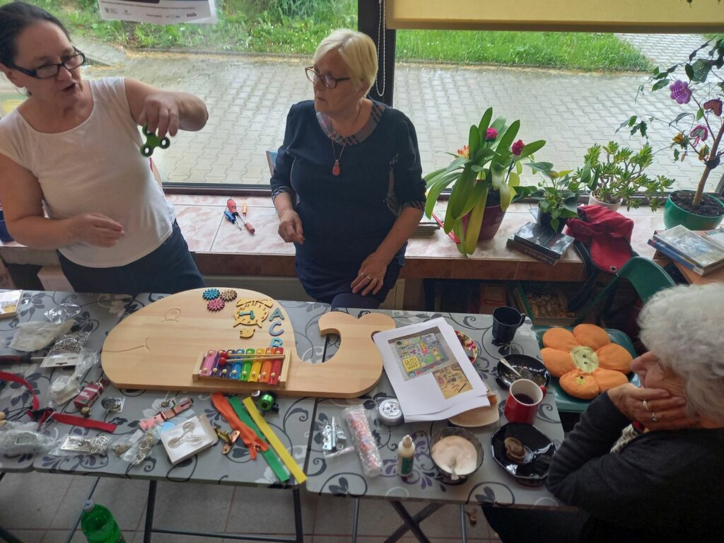 Przy stole stoja dwie panie z boku siedzi jedna pani