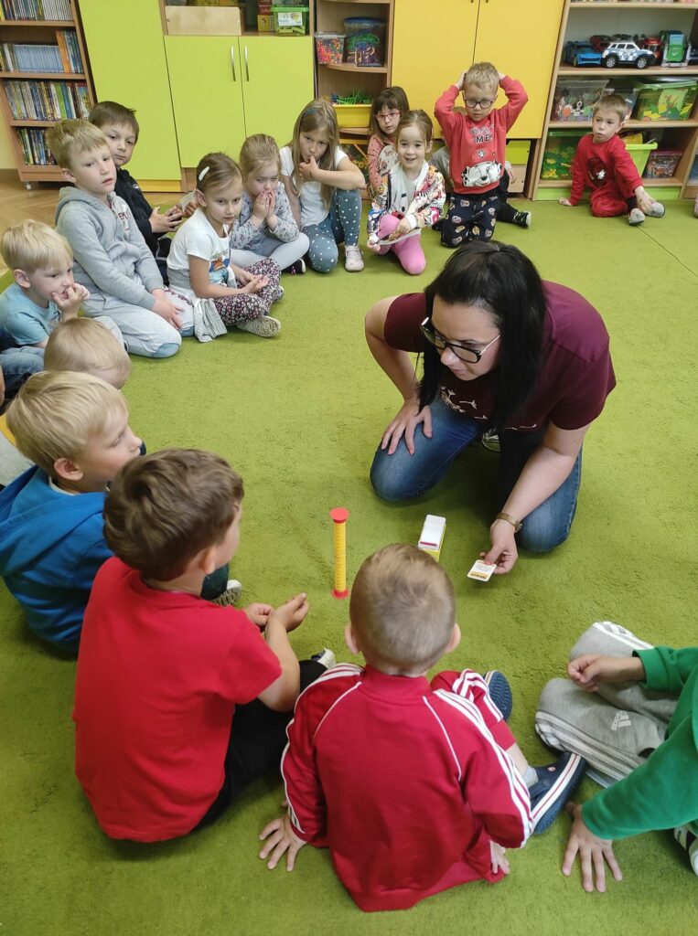 Na zdjęciu widać zielony dywan na którym siedzą dzieci a przed nimi klęczy bibliotekarka zadająca im zagadki,