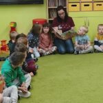 Na zdjęciu widać zielony dywan na którym siedzą dzieci oraz czytającą im bibliotekarkę.
