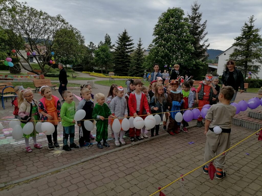 Na zdjęciu widoczna jest grupa przedszkolaków biorących udział w konkurencjach sportowych. Dzieci ubrane są w kolorowe stroje przedstawiające różne zwierzątka (pszczółki, motyle, żabki, wilki, zające, liski, bociany itp.)