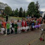 Na zdjęciu widoczna jest grupa przedszkolaków biorących udział w konkurencjach sportowych. Dzieci ubrane są w kolorowe stroje przedstawiające różne zwierzątka (pszczółki, motyle, żabki, wilki, zające, liski, bociany itp.)