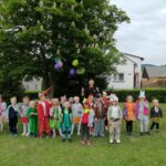 Na zdjęciu widoczna jest grupa przedszkolaków w kolorowych strojach przedstawiających różne zwierzątka (pszczółki, motyle, żabki, wilki, zające, liski, bociany itp.) pozujących do wspólnego zdjęcia z trenerem po zakończonych konkurencjach sportowych. W tle drzewa i domy.