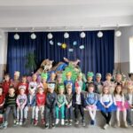 Na zdjęciu widoczna jest grupa 30 przedszkolaków 15 dziewczynek i 15 chłopców będących uczestnikami imprezy z okazji Dnia Dziecka. Dzieci ubrane są w kolorowe stroje przedstawiające różne zwierzęta lasów i łąk biedronki, misie, motyle, żaby, pszczółki, liski, zające,wilki, bociany. W tle dekoracja choinka, pagórki, sarenka, lis, strumyk, słońce i chmurki.