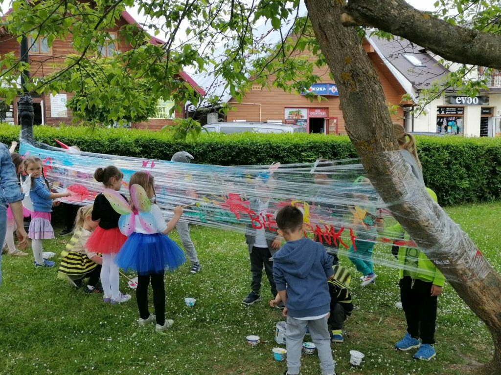 Na zdjęciu widoczna jest grupa przedszkolaków malujących na streczu rozciągniętym wokół drzew. Dzieci ubrane są w kolorowe stroje przedstawiające pszczółki, motylki, zajączki i żabki.