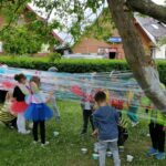 Na zdjęciu widoczna jest grupa przedszkolaków malujących na streczu rozciągniętym wokół drzew. Dzieci ubrane są w kolorowe stroje przedstawiające pszczółki, motylki, zajączki i żabki.