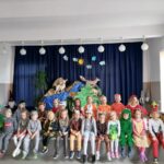 Na zdjęciu widoczna jest grupa 30 przedszkolaków 15 dziewczynek i 15 chłopców będących uczestnikami imprezy z okazji Dnia Dziecka. Dzieci ubrane są w kolorowe stroje przedstawiające różne zwierzęta lasów i łąk biedronki, misie, motyle, żaby, pszczółki, liski, zające,wilki, bociany. W tle dekoracja choinka, pagórki, sarenka, lis, strumyk, słońce i chmurki.