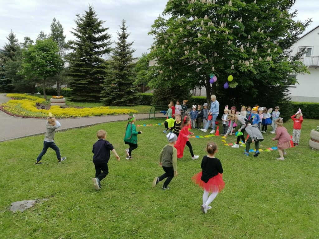 Na zdjęciu widoczna jest grupa przedszkolaków biorących udział w konkurencjach sportowych. Dzieci ubrane są w kolorowe stroje przedstawiające różne zwierzątka (pszczółki, motyle, żabki, wilki, zające, liski, bociany itp.)