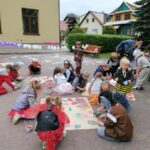 Na zdjęciu widoczna jest grupa przedszkolaków malujących farbami na rozłożonym szarym papierze. Dzieci ubrane są w kolorowe stroje (bociany, biedronki, jeże, wilki, liski, pszczółki itp.) W tle budynek WDK w Tyliczu oraz rozciągnięte na sznurku balony.
