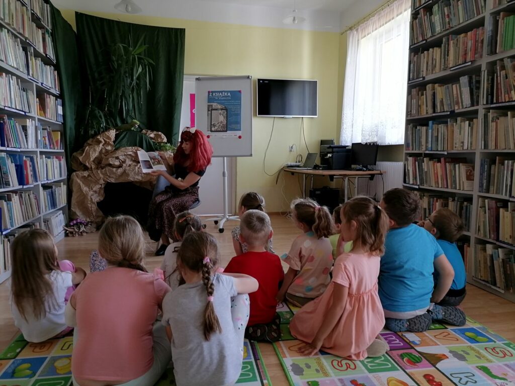 Na zdjęciu w sali bibliotecznej na podłodze siedzą dzieci. Przed nimi na krześle siedzi Pani bibliotekarka i czyta dzieciom bajeczkę.