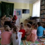 Na zdjęciu w sali bibliotecznej na podłodze siedzą dzieci. Przed nimi na krześle siedzi Pani bibliotekarka i czyta dzieciom bajeczkę.