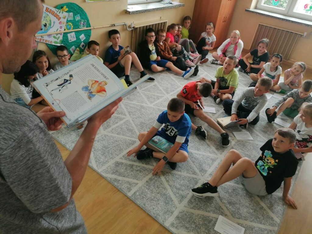 Cała grupa dzieci siedzi wygodnie na dywanie i słucha jak stojący przed nimi pan bibliotekarz czyta im książkę pt. „Guzik z mleka”.