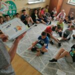 Cała grupa dzieci siedzi wygodnie na dywanie i słucha jak stojący przed nimi pan bibliotekarz czyta im książkę pt. „Guzik z mleka”.