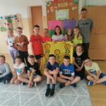 Pamiątkowe zdjęcie jednej z klas biorącej udział w spotkaniu. Uczniowie razem z panem bibliotekarzem stoją obok siebie, a pomiędzy nimi znajduje się żółta tarcza.